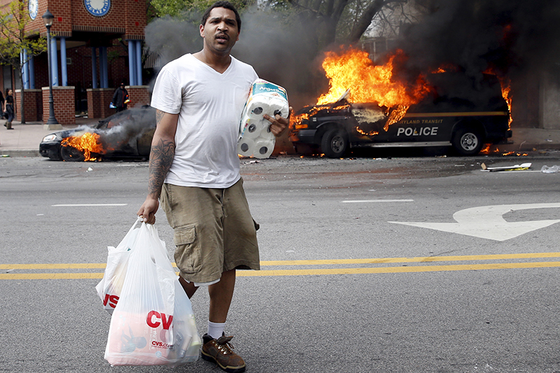 Человек с похищенными в магазине товарами проходит мимо горящих машин во время столкновений в Балтиморе