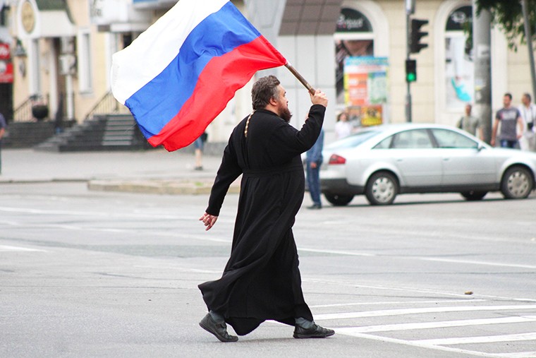 Православный священник идет с российским флагом по Луганску.
