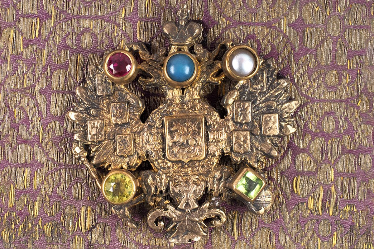 В верхней половине передней крышки помещена металлическая накладка в виде герба Российской империи. Фигура инкрустирована пятью ювелирными камнями (рубин, бирюза, жемчуг, демантоид и хризолит)