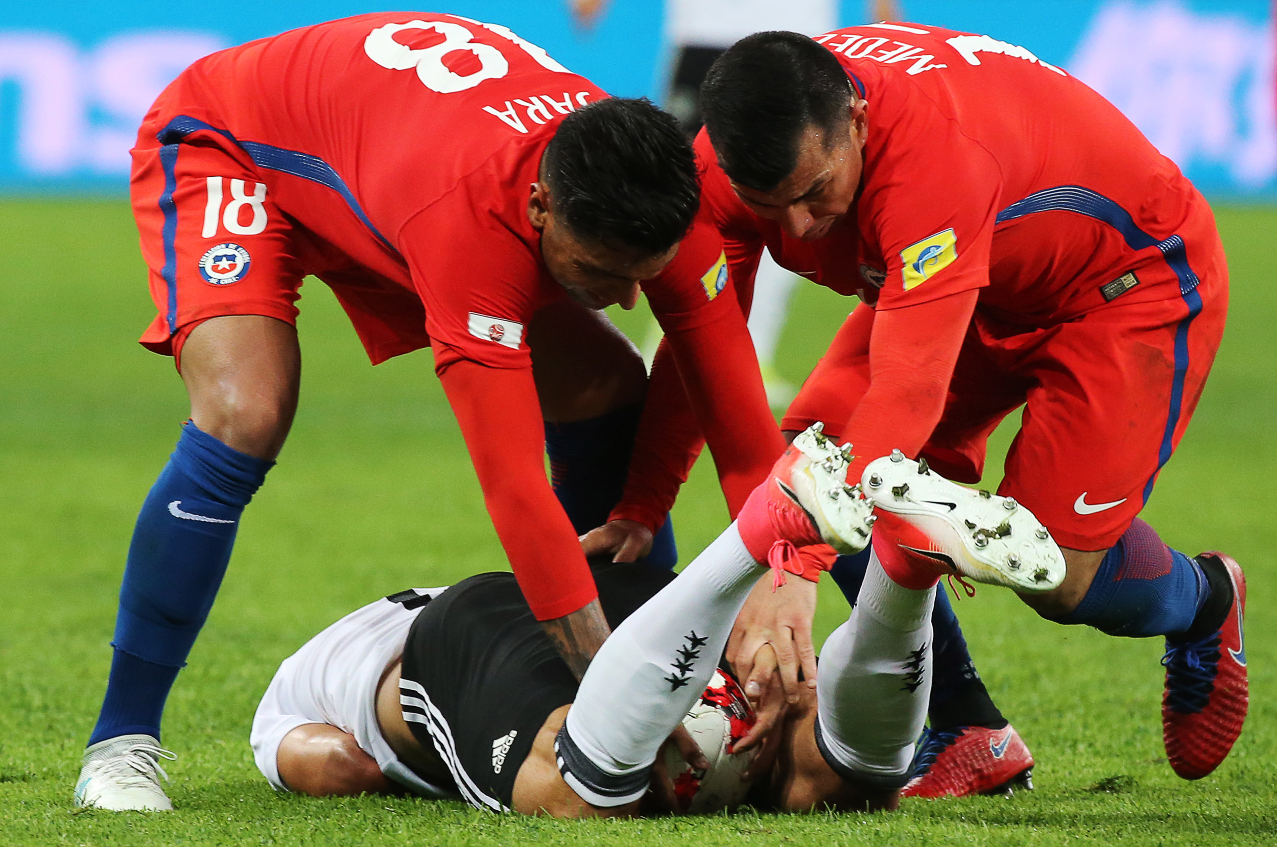 Игроки сборной Чили Гонсало Хара (№&nbsp;18) и Гари Медель пытаются поднять с газона немца Эмре Джана. С их точки зрения, соперник просто тянул время